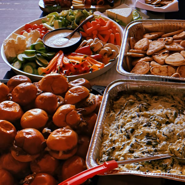 A buffet spread featuring mini burgers, a vegetable platter with dip, crackers, and a cheesy spinach dip.