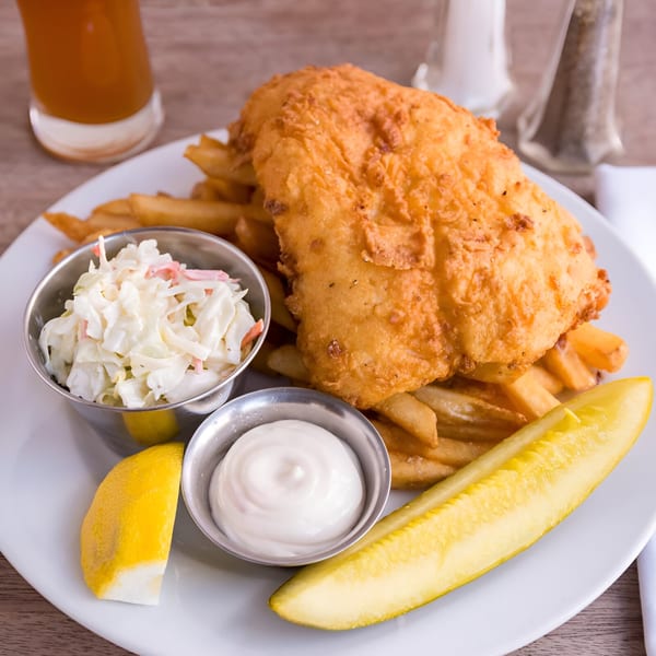 Golden-brown fish and chips with coleslaw, tartar sauce, lemon, and a pickle spear on a white plate.