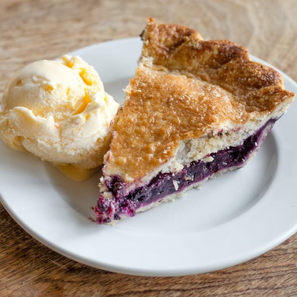 A slice of blueberry pie with a scoop of vanilla ice cream on a white plate.