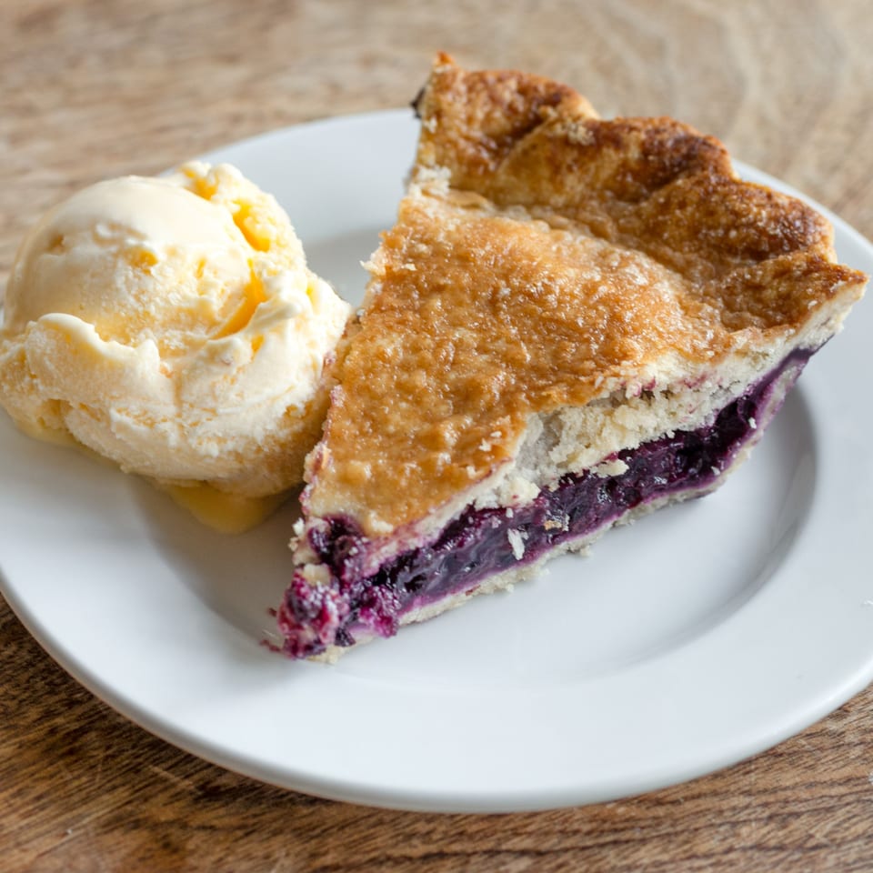 A slice of blueberry pie with a scoop of vanilla ice cream on a white plate.