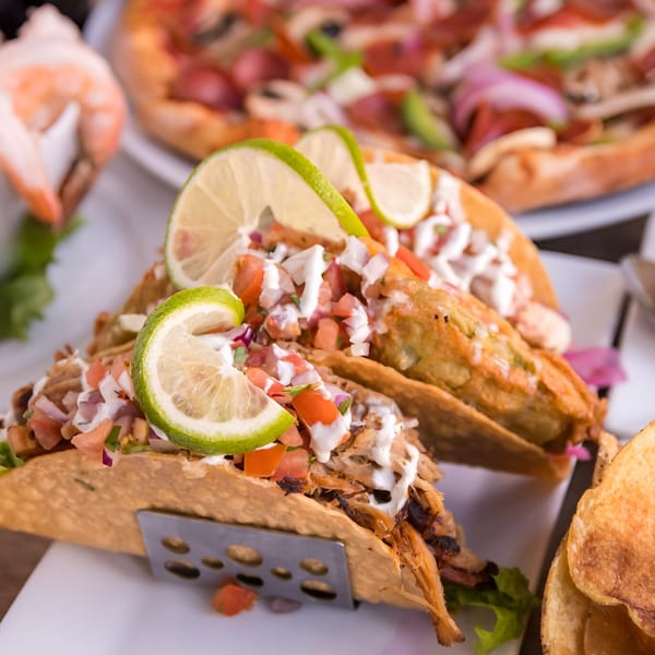 Two tacos with pico de gallo, sour cream, and lime wedges, with a pizza in the background.