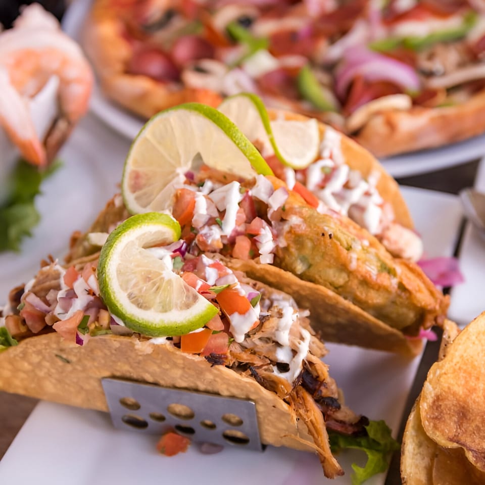 Two tacos with pico de gallo, sour cream, and lime wedges, with a pizza in the background.